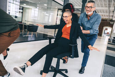 Group of businesspeople having fun during playtime. Cheerful group of colleagues pushing each other on chairs in a modern workplace. Team of businesspeople having fun and celebrating their success. - JLPSF07687