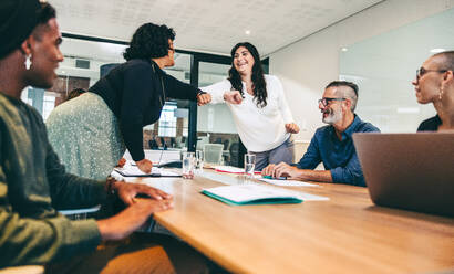 Female colleagues elbow bumping each other before a meeting in a boardroom. Group of happy businesspeople attending a briefing in a modern workplace. Modern businesspeople taking COVID-19 precautions. - JLPSF07706