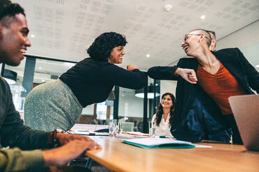 Lächelnde Geschäftsfrauen, die sich vor einer Sitzung in einem Sitzungssaal mit den Ellbogen stoßen. Eine Gruppe glücklicher Geschäftsleute, die an einer Besprechung an einem modernen Arbeitsplatz teilnehmen. Moderne Geschäftsleute, die COVID-19-Vorsichtsmaßnahmen treffen. - JLPSF07707