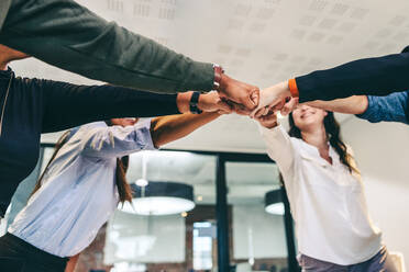 Kreative Geschäftsleute ballen ihre Fäuste, während sie an einem modernen Arbeitsplatz stehen. Verschiedene Gruppen von Geschäftsleuten lächeln fröhlich, während sie zusammen in einem Huddle stehen. - JLPSF07713