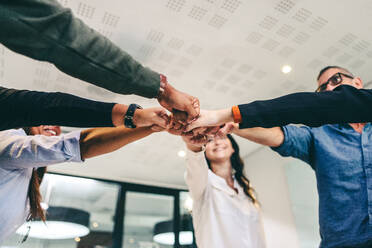 Moderne Geschäftsleute ballen ihre Fäuste, während sie an einem kreativen Arbeitsplatz stehen. Verschiedene Gruppen von Geschäftsleuten lächeln fröhlich, während sie zusammen in einem Huddle stehen. - JLPSF07714