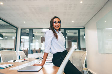 Happy businesswoman smiling at the camera in a boardroom. Mid-adult businesswoman leaning against a table in a modern workplace. Cheerful female entrepreneur wearing business casual in an office. - JLPSF07717