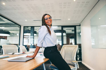 Businesswoman smiling cheerfully in a boardroom. Mid-adult businesswoman leaning against a table in a modern workplace. Cheerful female entrepreneur wearing business casual in a creative office. - JLPSF07718
