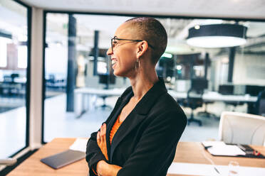 Successful female entrepreneur smiling cheerfully in a boardroom. Happy young businesswoman standing sideways with her arms crossed. Young businesswoman wearing business casual in a modern workplace. - JLPSF07720