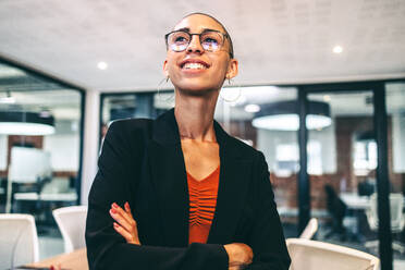 Glückliche junge Unternehmerin, die mit einem Lächeln im Gesicht in die Ferne schaut. Fröhliche junge Geschäftsfrau, die mit verschränkten Armen an einem modernen Arbeitsplatz steht. - JLPSF07721