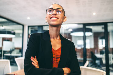 Looking forward to a bright future. Happy young female entrepreneur looking away with a smile on her face. Cheerful young businesswoman standing with her arms crossed in a modern workplace. - JLPSF07721