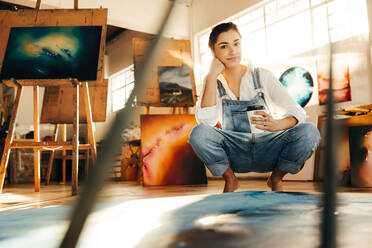 Junge freischaffende Malerin mit einer Tasse Kaffee in ihrem Atelier. Kreative junge Frau, die selbstbewusst in die Kamera schaut, während sie neben ihrem neuen Bild auf dem Boden hockt. - JLPSF07725