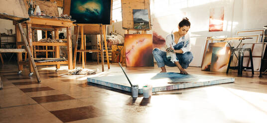 Eine Künstlerin malt ein blaues Bild auf einer Leinwand. Eine Malerin berührt ihr Bild, während sie in ihrem Atelier auf dem Boden hockt. Eine kreative junge Frau arbeitet an einem neuen Projekt. - JLPSF07727