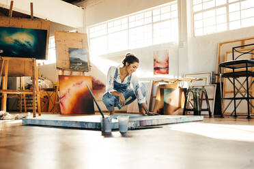 Fröhliche Künstlerin, die ein blaues Bild auf einer Leinwand malt. Kreative junge Frau, die glücklich lächelt, während sie ihr Bild auf dem Boden betrachtet. Malerin, die in ihrem Atelier an einem neuen Projekt arbeitet. - JLPSF07729