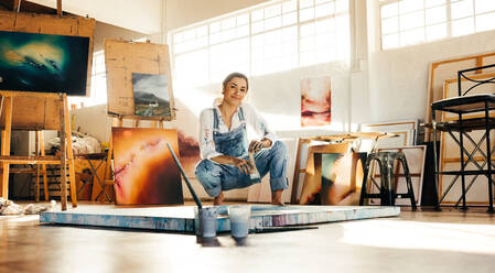 Creative painter squatting close to her painting in an art studio. Artistic young woman working on a new painting on the floor. Cheerful young artist looking at the camera while holding a paintbrush. - JLPSF07730