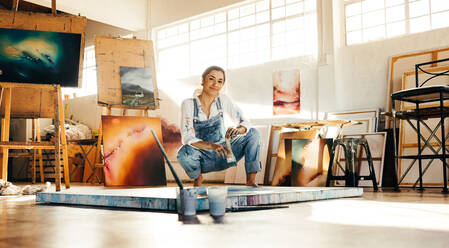 Kreative Malerin, die in einem Atelier neben ihrem Bild hockt. Künstlerische junge Frau, die auf dem Boden an einem neuen Bild arbeitet. Fröhliche junge Künstlerin, die in die Kamera schaut und einen Pinsel hält. - JLPSF07730
