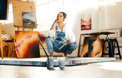 Junge Künstlerin, die auf dem Boden hockt und in ihrem Atelier arbeitet. Eine Malerin, die in die Kamera schaut und einen Pinsel hält. Eine kreative junge Frau, die ein neues Bild auf einer Leinwand malt. - JLPSF07733