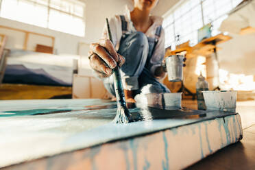 Unbekannte Künstlerin, die mit einem Pinsel auf einer großen Leinwand malt. Junge Malerin, die auf dem Boden ihres Ateliers arbeitet. Kreative junge Frau, die ein blaues Bild für ihr neues Kunstprojekt malt. - JLPSF07737