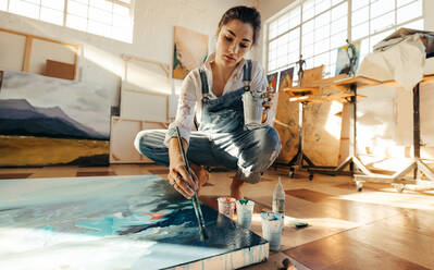 Female artist painting on a large canvas with a paintbrush. Imaginative young woman working on the floor of her art studio. Creative female painter making a blue artwork for her new project. - JLPSF07738