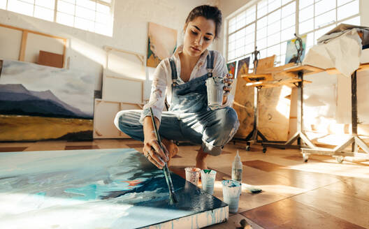 Eine Künstlerin malt mit einem Pinsel auf einer großen Leinwand. Eine phantasievolle junge Frau arbeitet auf dem Boden ihres Ateliers. Eine kreative Malerin fertigt ein blaues Kunstwerk für ihr neues Projekt an. - JLPSF07738