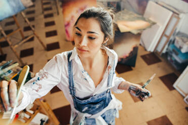 Young artist painting with a paintbrush in her studio. Creative young painter making a new artwork for her project. Female artist standing in front of a canvas in her atelier. - JLPSF07748