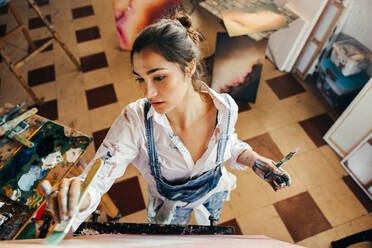 Creative young artist painting with a paintbrush in her studio. Imaginative young painter making a new artwork for her project. Female artist standing in front of a canvas in her atelier. - JLPSF07749