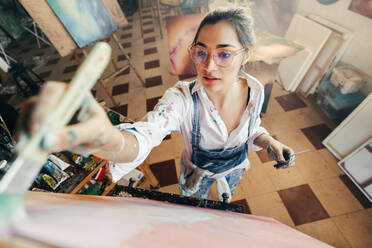 Künstlerin malt mit getönter Brille. Phantasievolle junge Malerin, die ein neues Kunstwerk für ihr Projekt anfertigt. Kreative Künstlerin vor einer Leinwand in ihrem Atelier. - JLPSF07750