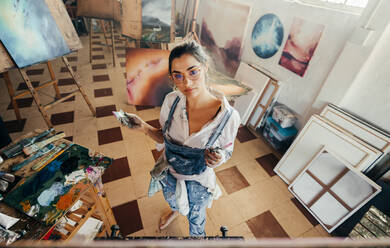 Blick von oben auf eine Malerin bei der Arbeit in ihrem Atelier. Einfallsreiche junge Künstlerin bei der Betrachtung ihres Bildes auf einer Leinwand. Kreative Künstlerin bei der Arbeit an einem neuen Kunstwerk für ihr Projekt. - JLPSF07751