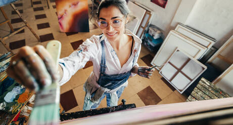 Fröhliche Künstlerin, die in ihrem Atelier an einer Leinwand malt. Blick von oben auf eine fantasievolle junge Malerin, die bei der Arbeit lächelt. Kreative Künstlerin, die ein neues Kunstwerk für ihr Projekt erstellt. - JLPSF07752