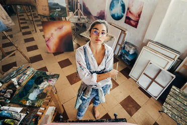 High angle view of an artist looking at the camera. Imaginative young painter standing in front of a canvas with her arms crossed. Creative young woman working on a new artwork for her project. - JLPSF07753