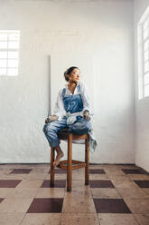 Kontemplative Künstlerin, die auf einem Stuhl in ihrem Atelier sitzt. Junge Malerin, die vor einer leeren Leinwand sitzt und über neue Ideen nachdenkt. Kreative junge Frau, die ein neues Kunstprojekt beginnt. - JLPSF07757