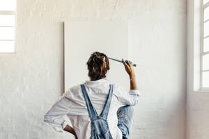 Contemplating creative ideas for a new project. Rearview of a young painter looking thoughtful while sitting in front of a blank canvas. Creative female artist holding a paintbrush to her head. - JLPSF07758