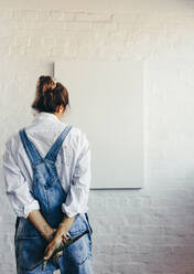 Nachdenkliche Malerin, die in ihrem Atelier mit dem Pinsel auf eine leere Leinwand blickt. Rückansicht einer fantasievollen jungen Künstlerin, die über kreative Ideen für ihr neues Projekt nachdenkt. - JLPSF07760