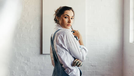 Confident female painter starting a new project in her studio. Female artist looking at the camera while standing in front of a blank canvas. Creative young woman holding a paintbrush. - JLPSF07763