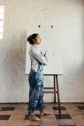 Female artist thinking of new creative ideas in her studio. Pensive young painter looking away while standing in front of a blank canvas. Imaginative young woman starting a new art project. - JLPSF07764