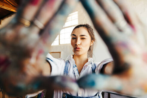 Female artist framing her face with her hands. Carefree female painter winking at the camera while standing in her art studio. Creative young woman making a frame with her colour painted hands. - JLPSF07767