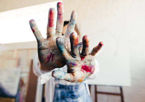 Eine Malerin, die mit ihren bunt bemalten Händen die Kamera von ihrem Gesicht abschirmt. Eine unerkennbare Künstlerin, die in ihrem Atelier steht, mit ihrer leeren Leinwand im Hintergrund. - JLPSF07769