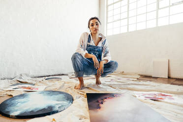 Talentierte Malerin, die in der Mitte ihrer Bilder hockt. Selbstbewusste junge Künstlerin, die in ihrer Kunstgalerie in die Kamera schaut. Kreative junge Frau, die eine Mappe mit ihren Kunstwerken zeigt. - JLPSF07773