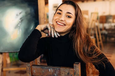 Glückliche professionelle Malerin, die in ihrem Atelier in die Kamera lächelt. Erfolgreiche Künstlerin, die auf einem Stuhl in ihrem Atelier sitzt, mit ihren Kunstwerken im Hintergrund. - JLPSF07787