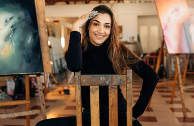 Erfolgreiche Malerin, die in ihrem Atelier in die Kamera lächelt. Glückliche professionelle Künstlerin, die auf einem Stuhl in ihrem Atelier sitzt, mit ihren Kunstwerken im Hintergrund. - JLPSF07788