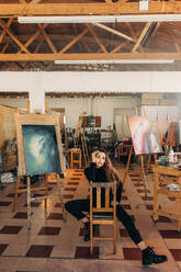 Selbstbewusste Künstlerin, die auf einem Stuhl in ihrem Atelier sitzt. Junge professionelle Malerin, die in die Kamera schaut, während sie mit ihrem Kunstwerk hinter sich sitzt. - JLPSF07789