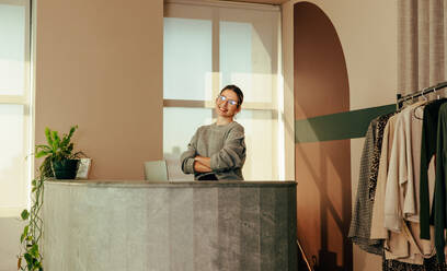 Female entrepreneur smiling at the camera while standing behind the counter in her clothing store. Successful small business owner working at the front desk in her fashion shop. - JLPSF07802