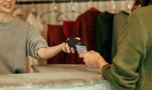 Unbekannte Kundin, die an der Kasse mit einer Kreditkarte bezahlt. Junge Frau, die eine Kreditkarte auf einem kontaktlosen Kartenlesegerät abtippt. Frau, die in einem Bekleidungsgeschäft Kleidung kauft. - JLPSF07806