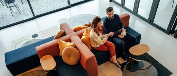 High angle view of two businesspeople working in an office lobby. Two modern businesspeople using a digital tablet while having a discussion. Two young entrepreneurs collaborating on a new project. - JLPSF07838