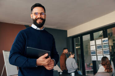 Successful businessman smiling at the camera while holding a digital tablet. Happy young businessman standing in a modern boardroom with his colleagues in the background. - JLPSF07857