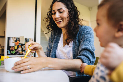 Maternity leave. Mother feeding her hungry baby sitting on high chair at home. Caring woman feeding her kid at home. - JLPSF07869