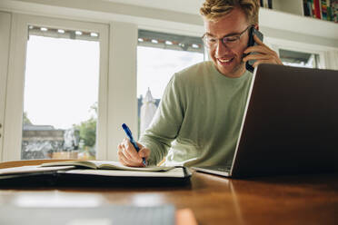 Man sitting at table with laptop talking on phone and writing in a book. Man working from home. - JLPSF07874