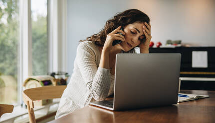 Woman feeling tired while working from home. Woman looking stressed sitting at table with laptop and talking on mobile phone. - JLPSF07876