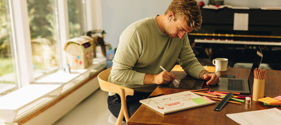 Freiberuflicher Illustrator, der von seinem Büro zu Hause aus arbeitet. Ein Mann sitzt am Tisch mit einem Laptop und zeichnet mit einem Stift auf seinem Grafiktablett. - JLPSF07878