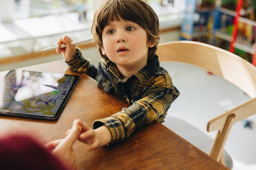 Cute boy sitting at table with digital tablet and looking at her mother. Boy learning to draw in a digital tablet at home. - JLPSF07882