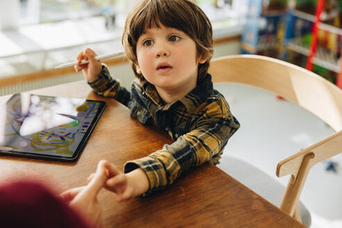 Süßer Junge sitzt am Tisch mit digitalem Tablet und schaut seine Mutter an. Junge lernt zu Hause mit einem digitalen Tablet zu zeichnen. - JLPSF07882