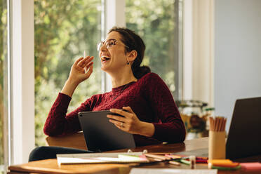 Cheerful designer sitting at home with digital tablet. Woman looking away and laughing while making illustrations on tablet computer. - JLPSF07900