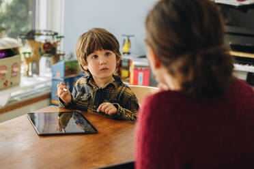 Ein Kind benutzt ein digitales Tablet und schaut seine Mutter an, die vorne am Tisch sitzt. Ein Junge lernt, auf einem Grafiktablett zu zeichnen, während seine Mutter daneben sitzt. - JLPSF07909