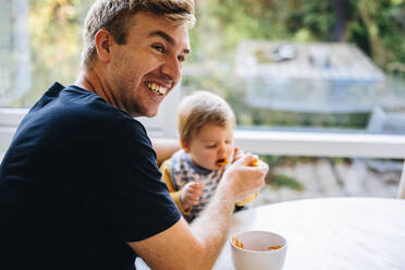 Lächelnder Mann, der sein Baby mit einem Löffel füttert, Vater, der sich um sein Baby kümmert, während er im Vaterschaftsurlaub ist. - JLPSF07915