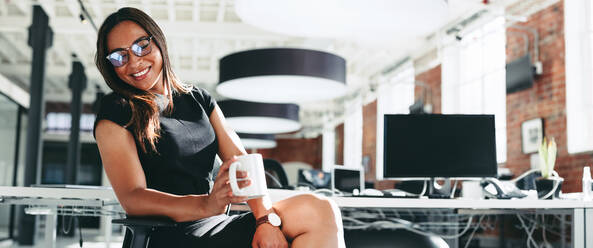 Enjoying a coffee break in the office. Happy young businesswoman smiling cheerfully while holding a cup of coffee. Young businesswoman sitting alone in a modern workplace. - JLPSF07928